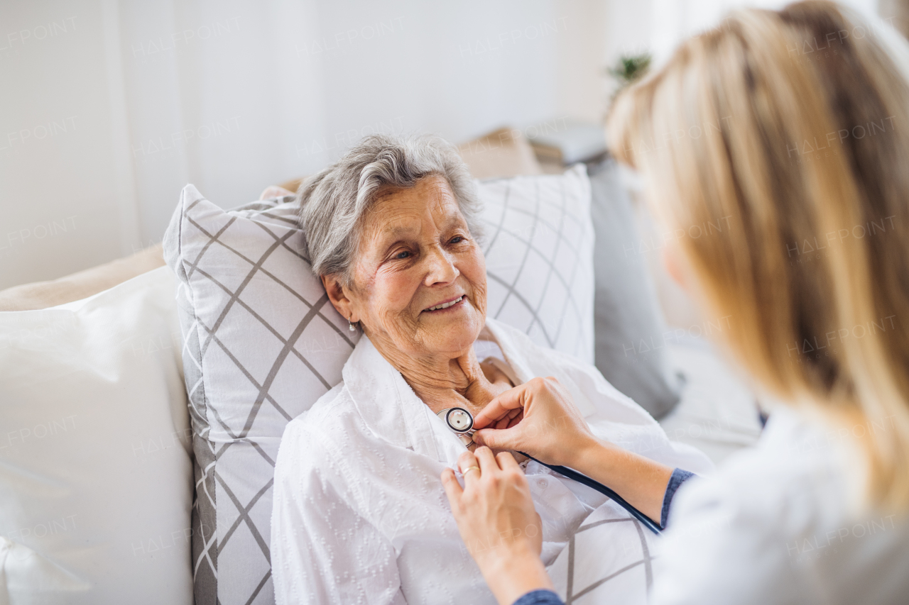 A young health visitor examining a sick senior woman lying in bed at home with stethoscope.