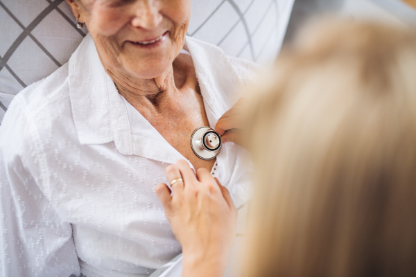 A midsection of young health visitor examining a sick senior woman lying in bed at home with stethoscope.