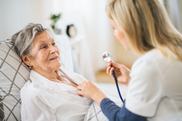 A young health visitor examining a sick senior woman lying in bed at home with stethoscope.