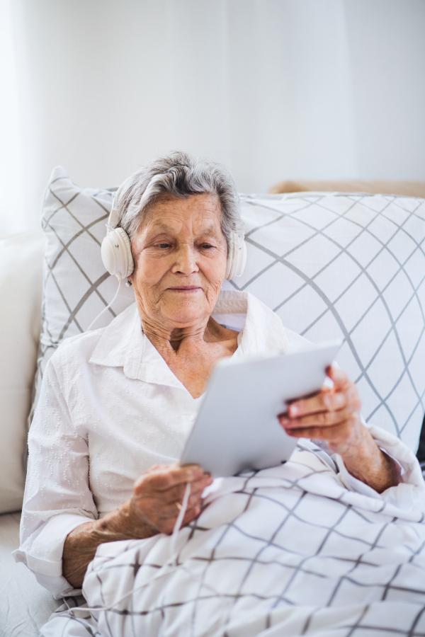 A sick senior woman with headphones and tablet lying in bed at home or in hospital, listening to music.