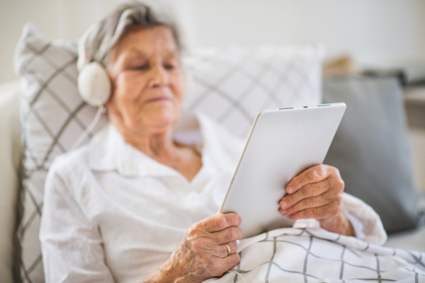 A sick senior woman with headphones and tablet lying in bed at home or in hospital, listening to music.