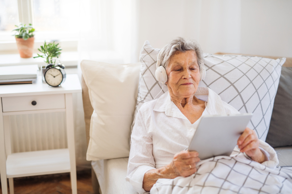 A sick senior woman with headphones and tablet lying in bed at home or in hospital, listening to music.