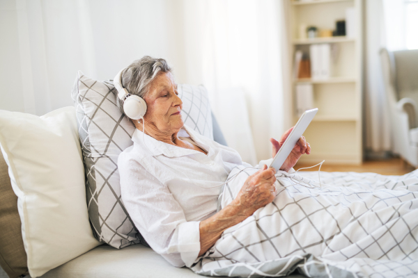 A sick senior woman with headphones and tablet lying in bed at home or in hospital, listening to music.