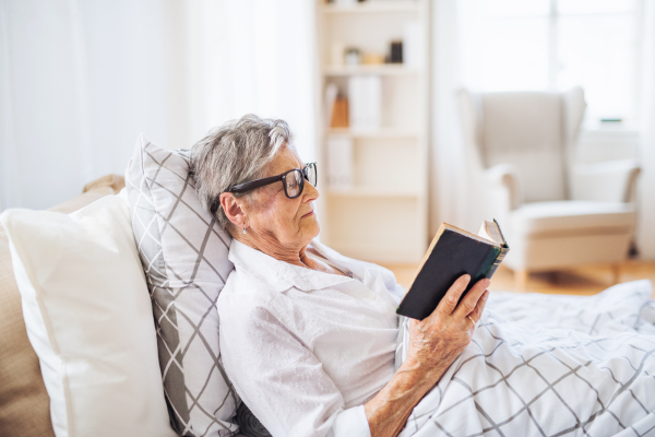 A sick senior woman with eyeglasses lying in bed at home or in hospital, reading bible book.