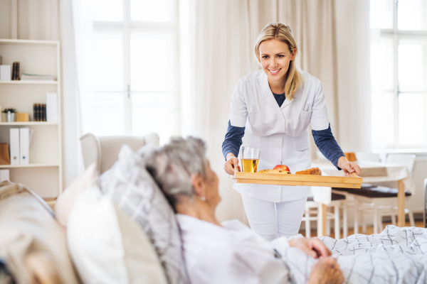 A young health visitor bringing breakfast to a sick senior woman lying in bed at home.