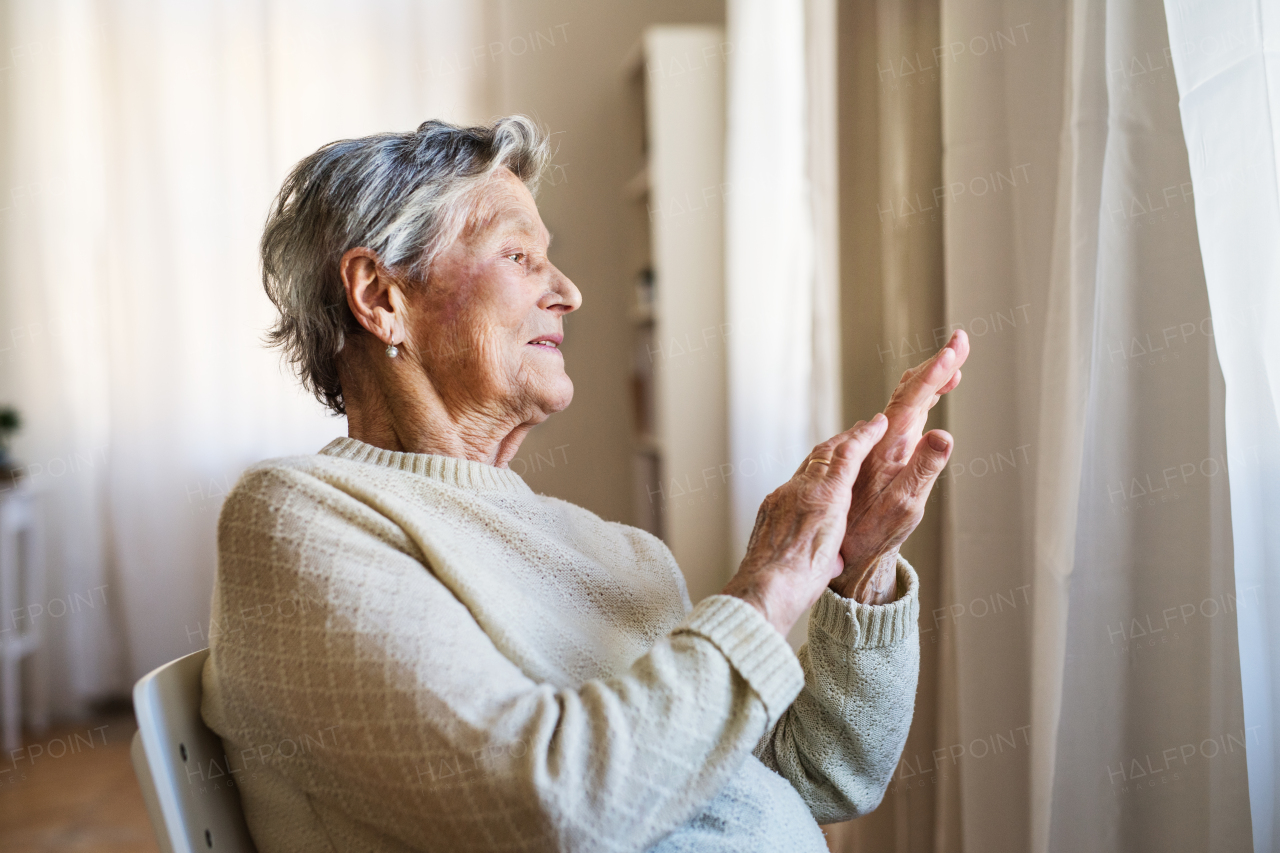 A portrait of a senior woman sitting on a chair at home, looking out of a window.