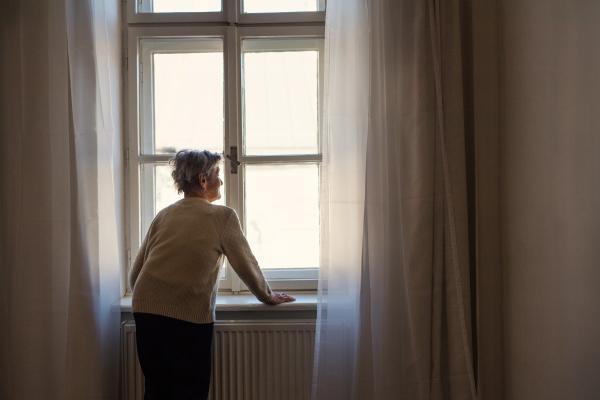 A rear view of a senior woman standing at home, looking out of a window. Copy space.