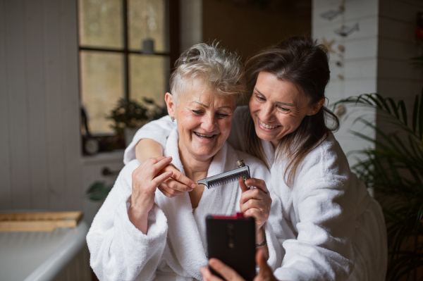 A happy senior mother in bathrobe with adult daughter taking selfie indoors at home, selfcare concept.