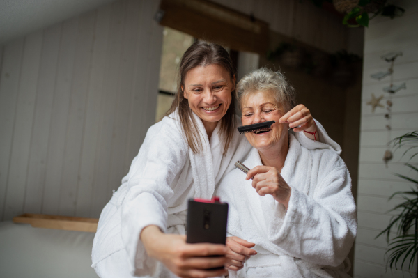 A happy senior mother in bathrobe with adult daughter taking selfie indoors at home, selfcare concept.