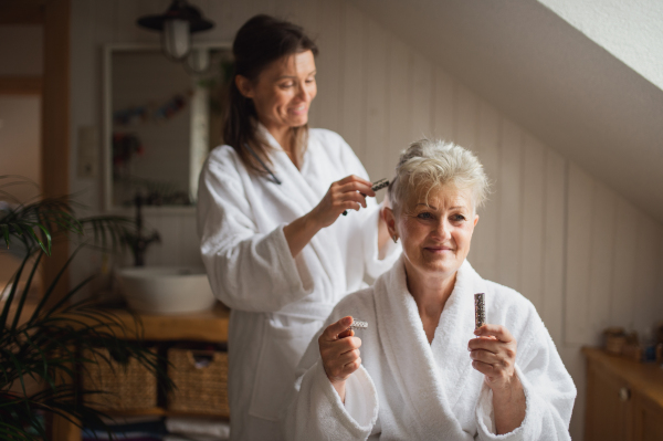 A happy senior mother in bathrobe with adult daughter using hair curlers indoors at home, selfcare concept.