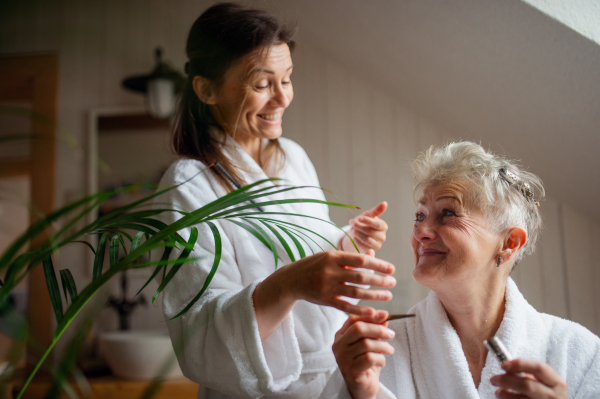 A happy senior mother in bathrobe with adult daughter indoors at home, selfcare concept.