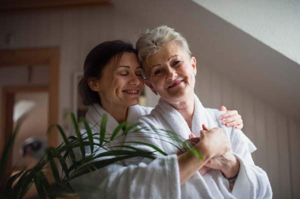 A happy senior mother in bathrobe with adult daughter hugging indoors at home, selfcare concept.