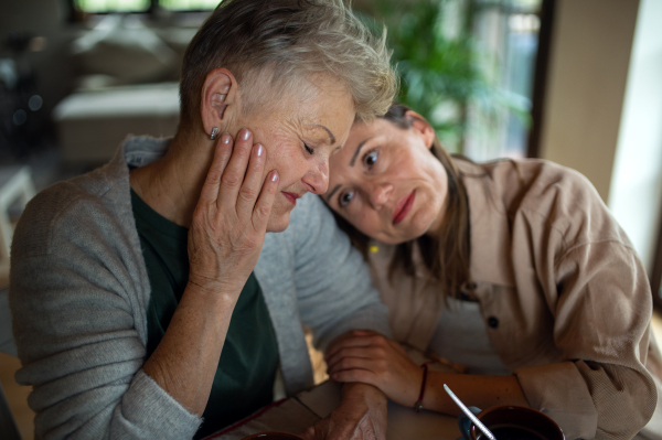 A portrait of sad senior mother with adult daughter indoors at home, hugging.