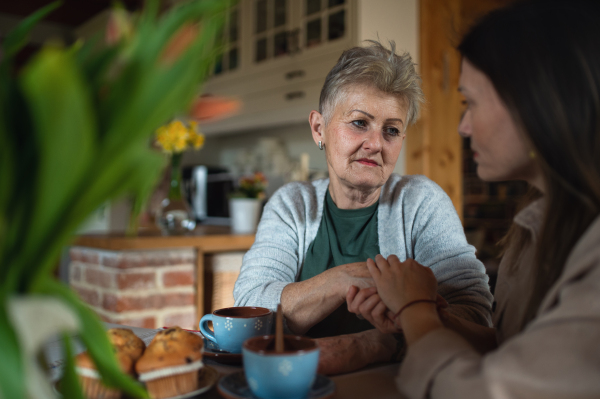 A sad senior mother having tea with adult daughter indoors at home, talking.