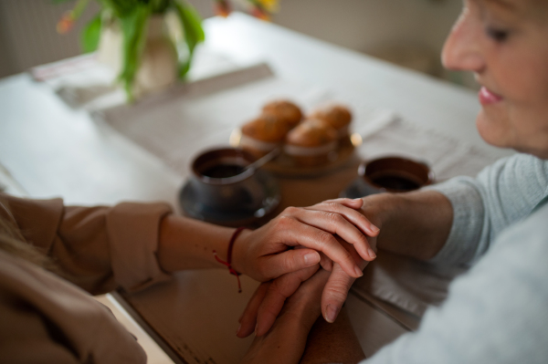 A close up of senior mother holding hands with adult daughter indoors at home, having coffee.