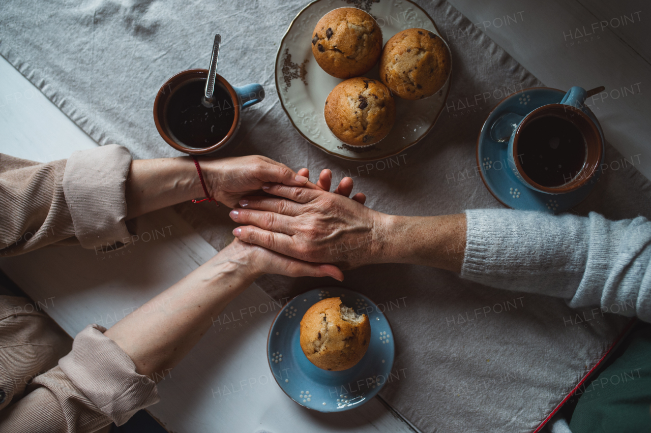 A top view of senior mother having coffee with adult daughter indoors at home, holding hands.