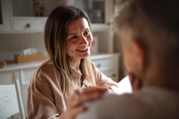 A happy adult daughter having tea with unrecognizable senior mother indoors at home, holding her shoulder.