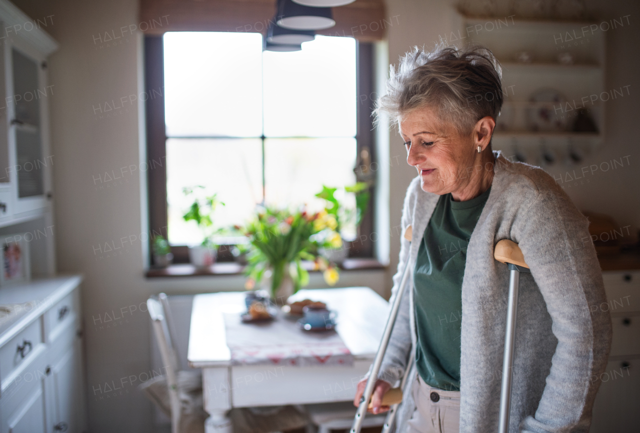 A senior woman with crutches indoors at home, walking in kitchen.