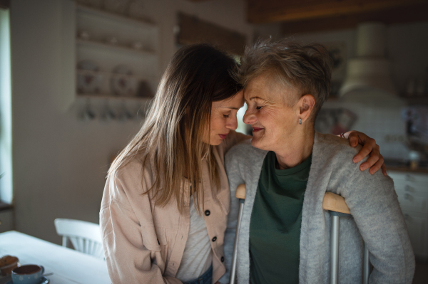 Happy adult daughter visiting ill senior mother with crutches indoors at home, hugging.