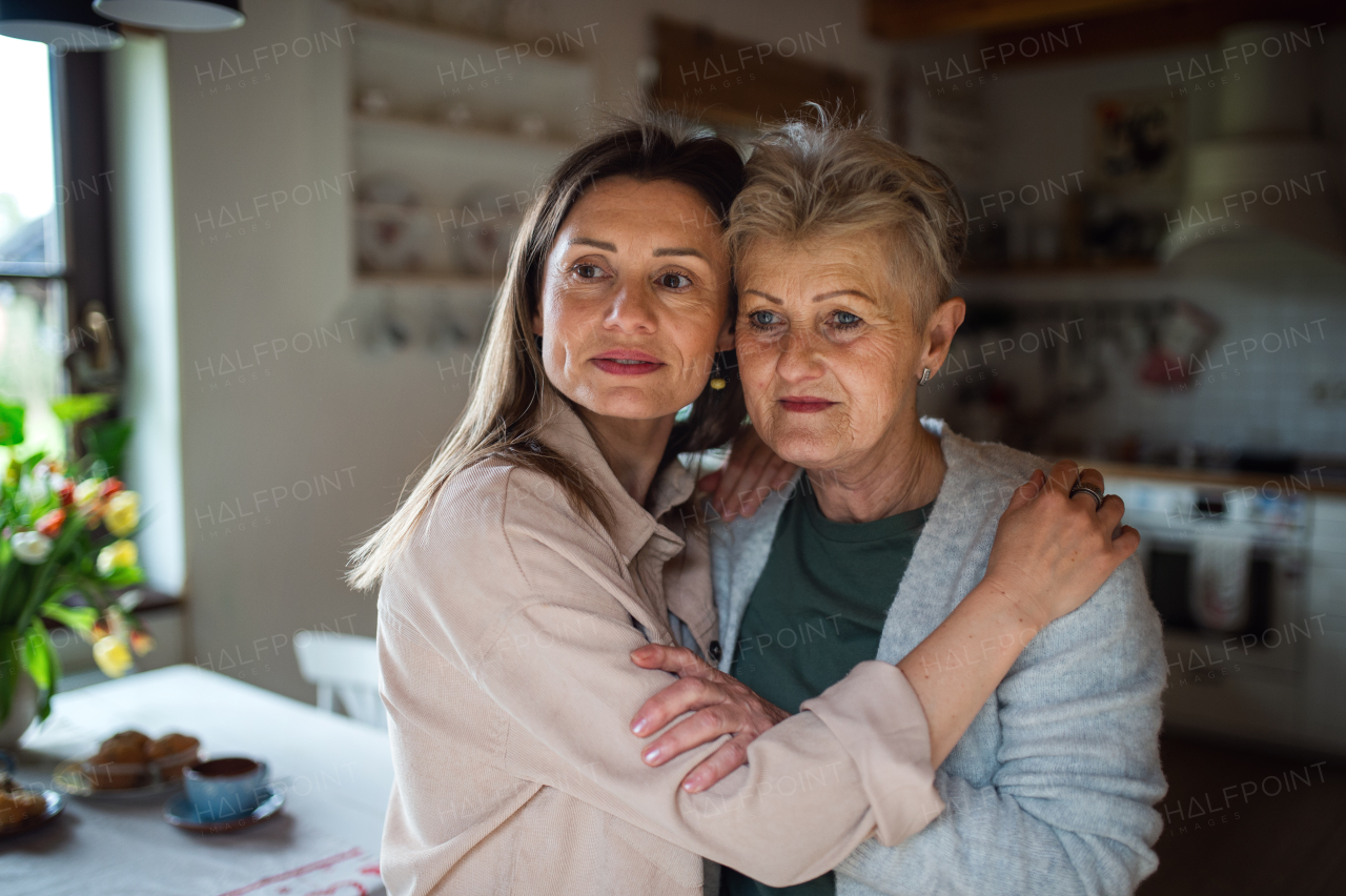 A portrait of senior mother hugging with adult daughter indoors at home, looking aside.