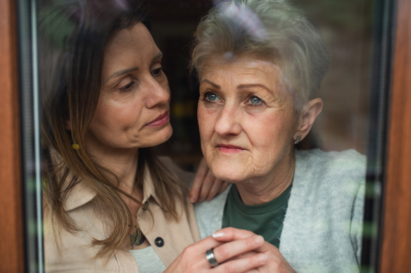 A close-up portrait of sad senior mother with adult daughter indoors at home, hugging.