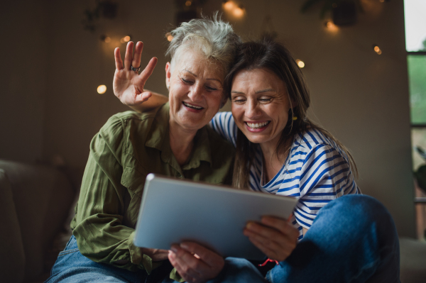 A portrait of happy senior mother with adult daughter using tablet indoors at home, video call concept.