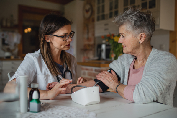 A healthcare worker or caregiver visiting senior woman indoors at home, measuring blood pressure.