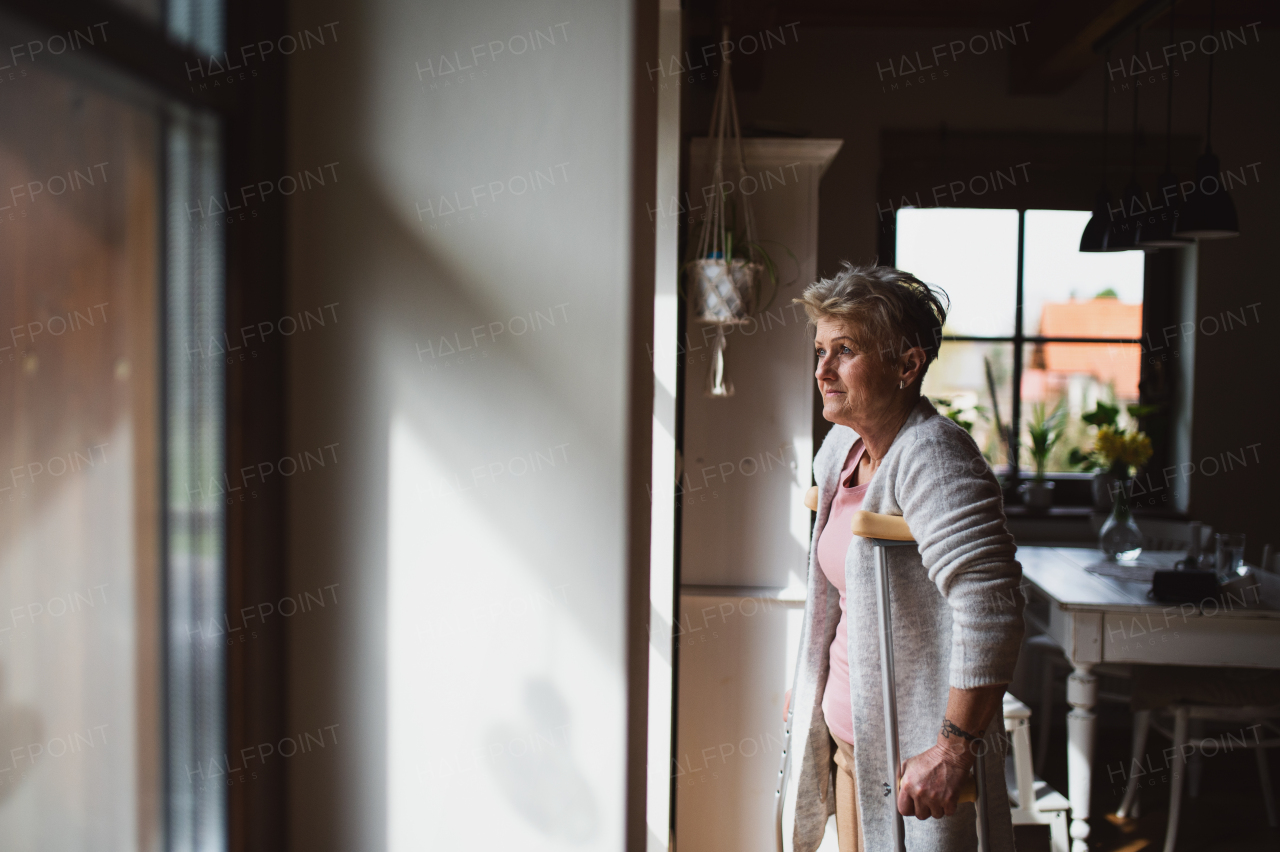 A sad senior woman with crutches indoors at home, looking out through window.