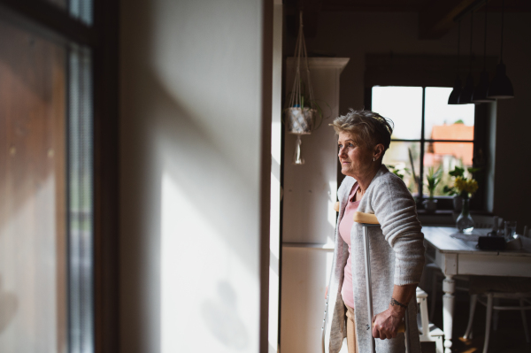 A sad senior woman with crutches indoors at home, looking out through window.