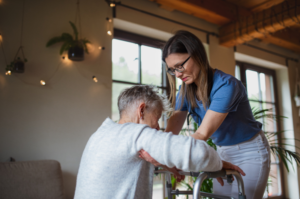 A healthcare worker or caregiver visiting senior woman indoors at home, helping her to walk.