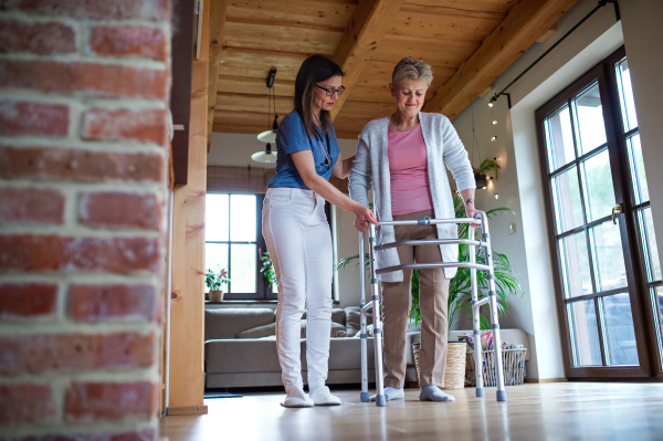 A healthcare worker or caregiver visiting senior woman indoors at home, helping her to walk.