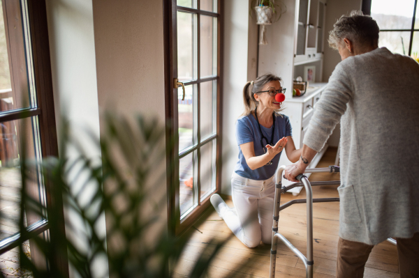 A healthcare worker or caregiver with red nose visiting senior woman indoors at home, having fun.