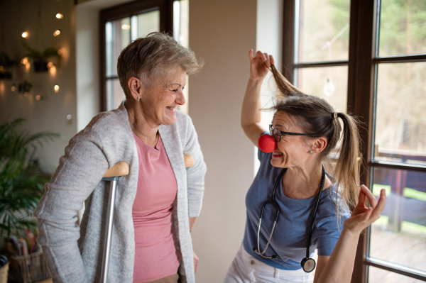 A healthcare worker or caregiver with red nose visiting senior woman indoors at home, having fun.