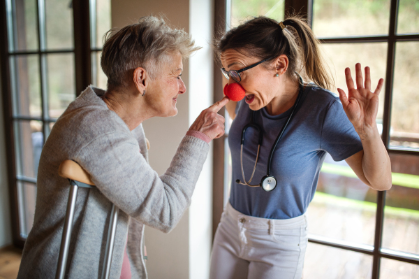 A healthcare worker or caregiver with red nose visiting senior woman indoors at home, having fun.