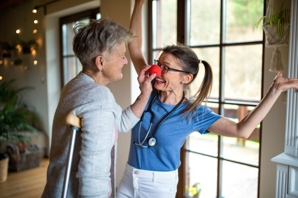 A healthcare worker or caregiver with red nose visiting senior woman indoors at home, having fun.