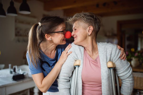 A healthcare worker or caregiver with red nose visiting senior woman indoors at home, having fun.