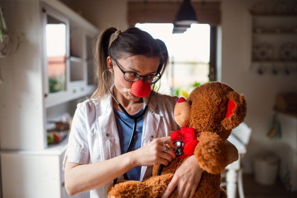 Portrait of a funny female doctor with red clown nose holding teddy bear.