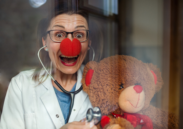 A healthcare worker or caregiver with red nose and teddy bear looking at camera inddors, shot through glass.