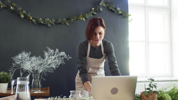 A young creative woman arranging flowers in a flower shop, using laptop. A startup of florist business.