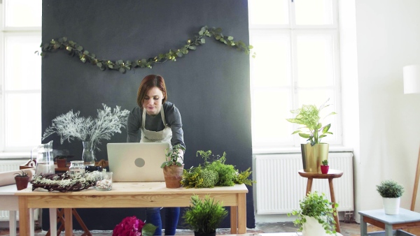 A young creative woman arranging flowers in a flower shop, using laptop. A startup of florist business.