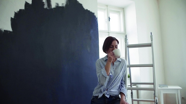 A young woman painting wall black, drinking coffee. A startup of small business. Slow motion.