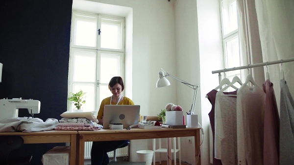 Young creative woman with laptop working in a studio, startup of small tailoring business. Slow motion.