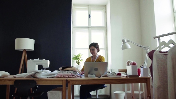 Young creative woman with laptop working in a studio, startup of small tailoring business. Slow motion.