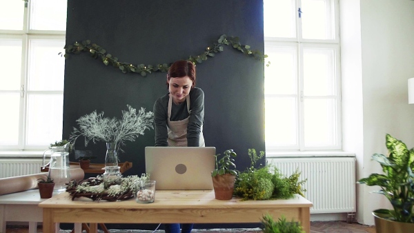 A young creative woman arranging flowers in a flower shop, using laptop. A startup of florist business. Slow motion.