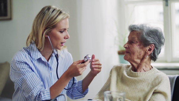 A young health visitor examining a senior woman with a stethoscope at home.