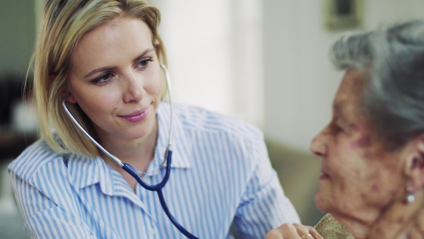 A young health visitor examining a senior woman with a stethoscope at home.