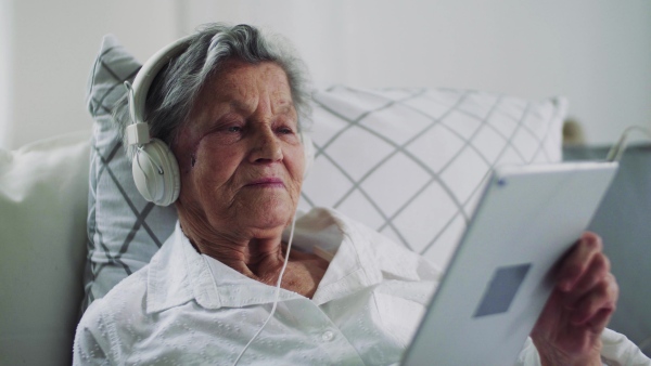 A sick senior woman with headphones and tablet lying in bed at home or in hospital, listening to music.