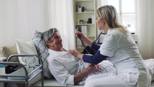 A young health visitor examining a sick senior woman lying in bed at home with stethoscope.