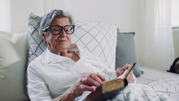 Sick senior woman with glasses lying in bed at home or in hospital, reading bible book.