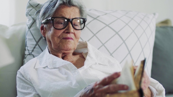 Sick senior woman with glasses lying in bed at home or in hospital, reading bible book.